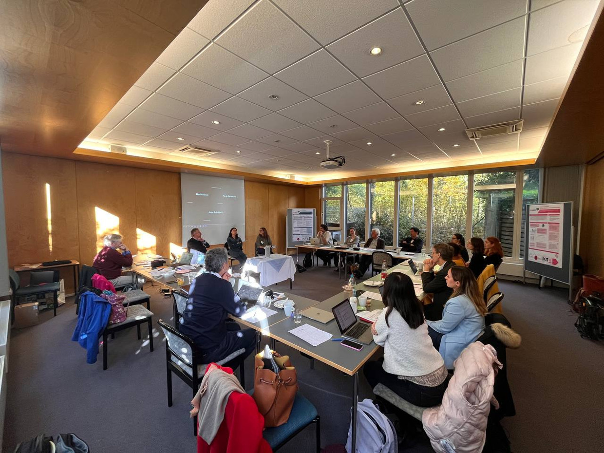 Anna Klassen Personen sitzen an Tischen, die zu einem U geformt sind. Drei Personen sitzen am offenen Ende des U auf Stühlen. Hinter ihnen ist eine Leinwand mit Namen zu sehen, die die online Teilnehmer*innen am Workshop zeigt.