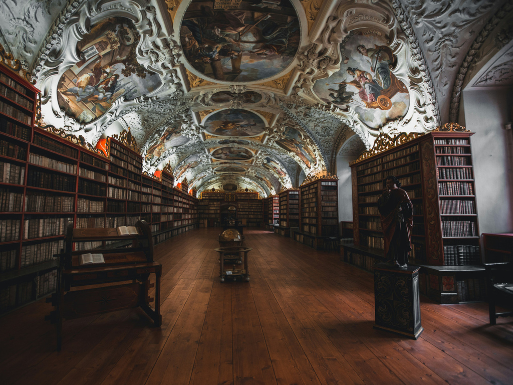 Eine historische Bibliothek mit Stock und Gemälden an der Decke, Fußboden, Regale und die Bücher sind dunkel gehalten, die Decke hell.